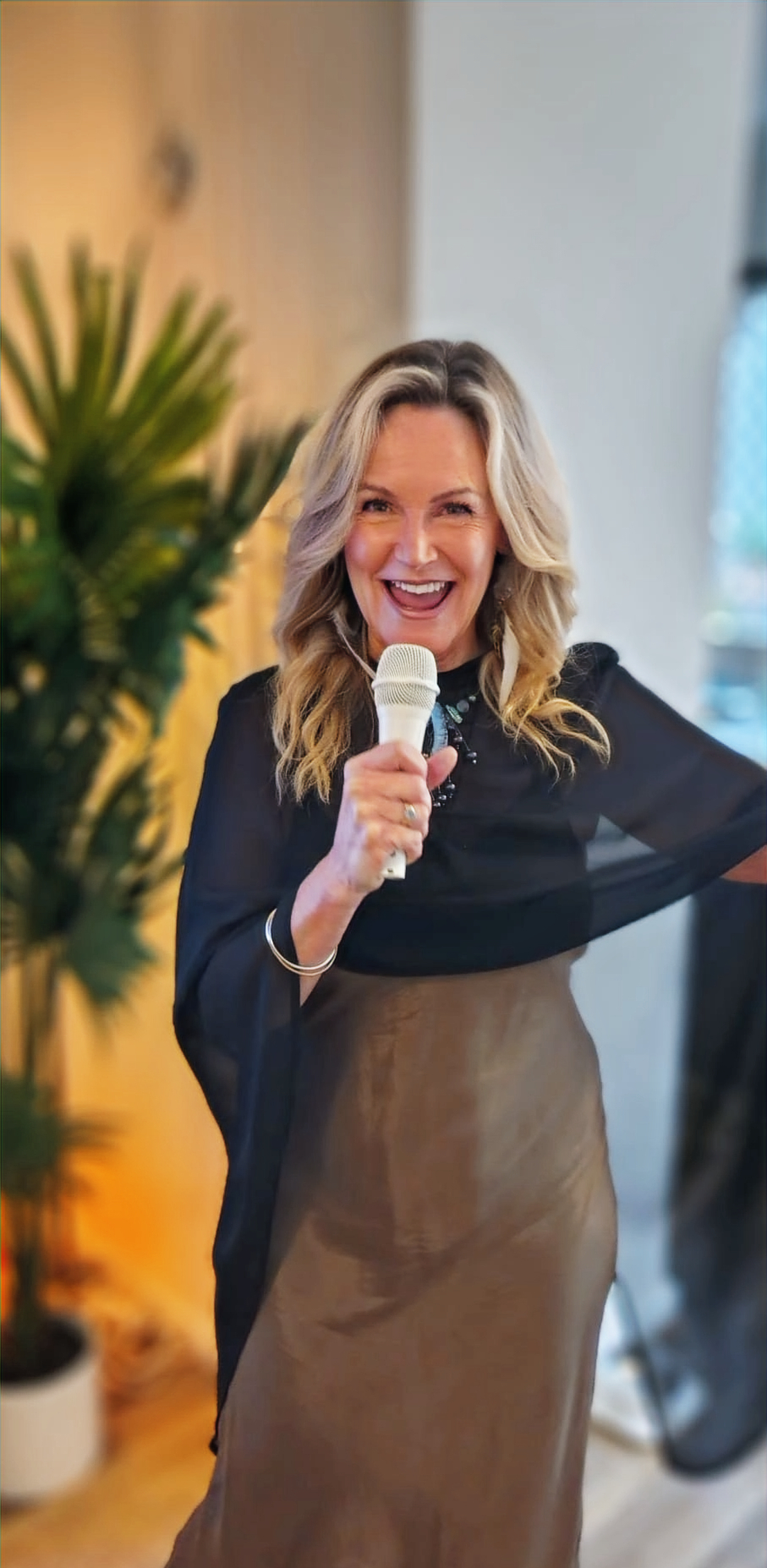 Krissy, a blonde woman in a black dress, smiles holding a microphone near a green plant at her wedding.