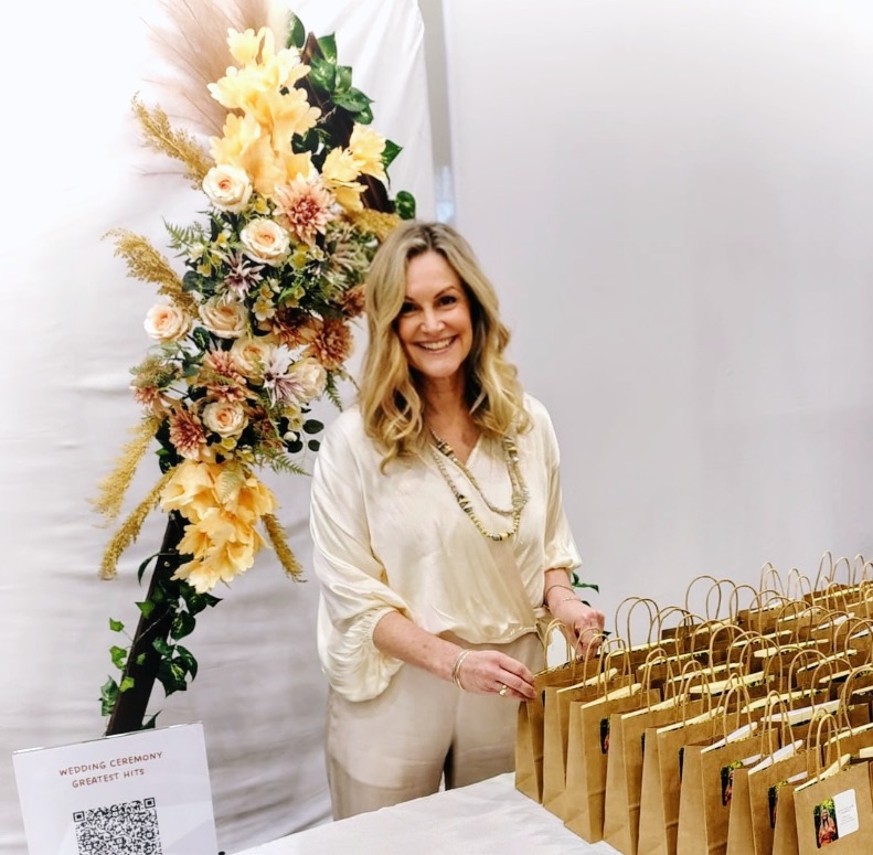 Krissy stands by a floral display and brown paper gift bags with a QR code sign at the loveiam wedding.