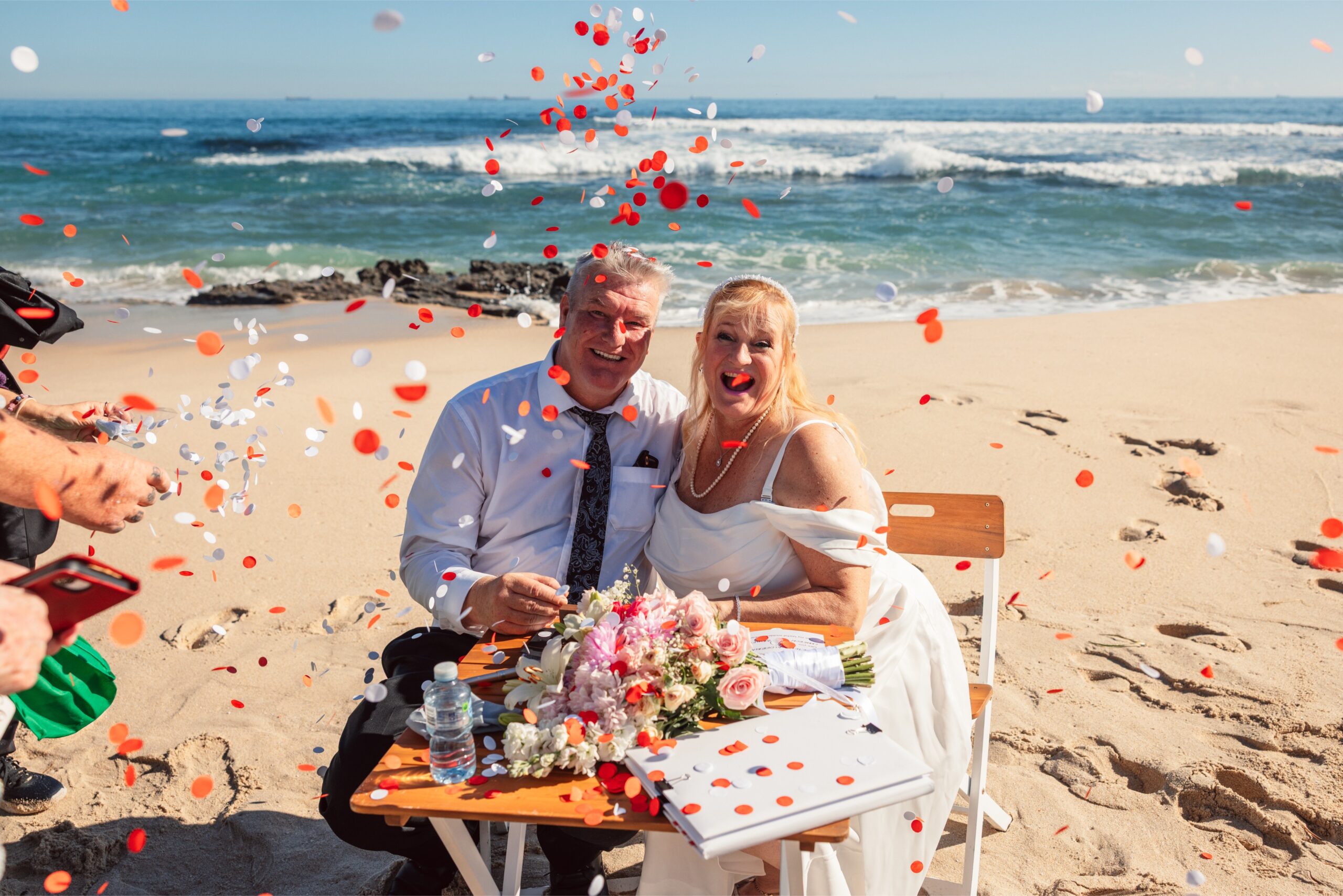 A couple in wedding attire smiles at a flower-decorated beach table as red and white confetti falls around them.